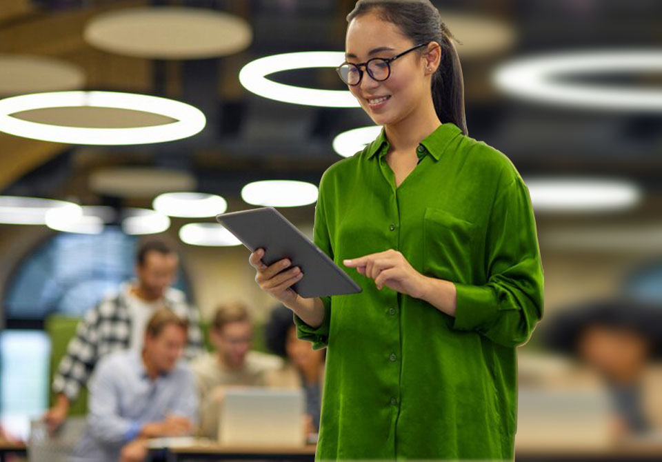 Garota de camisa verde sorrindo e vendo algo no tablet, ao fundo pessoas trabalhando